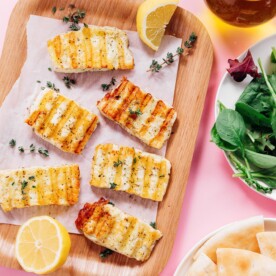 Grilled halloumi slices with herbs rest on a wooden tray, garnished with lemon wedges and thyme. This grilled halloumi is served alongside pita bread, fresh salad, and a drizzle of olive oil.