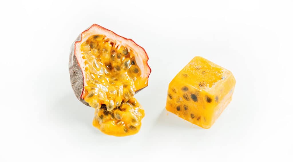 A cut open passion fruit with the filling oozing out next to a frozen passion fruit cube.