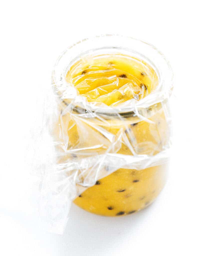 Passion fruit curd in a small glass jar covered with plastic wrap.