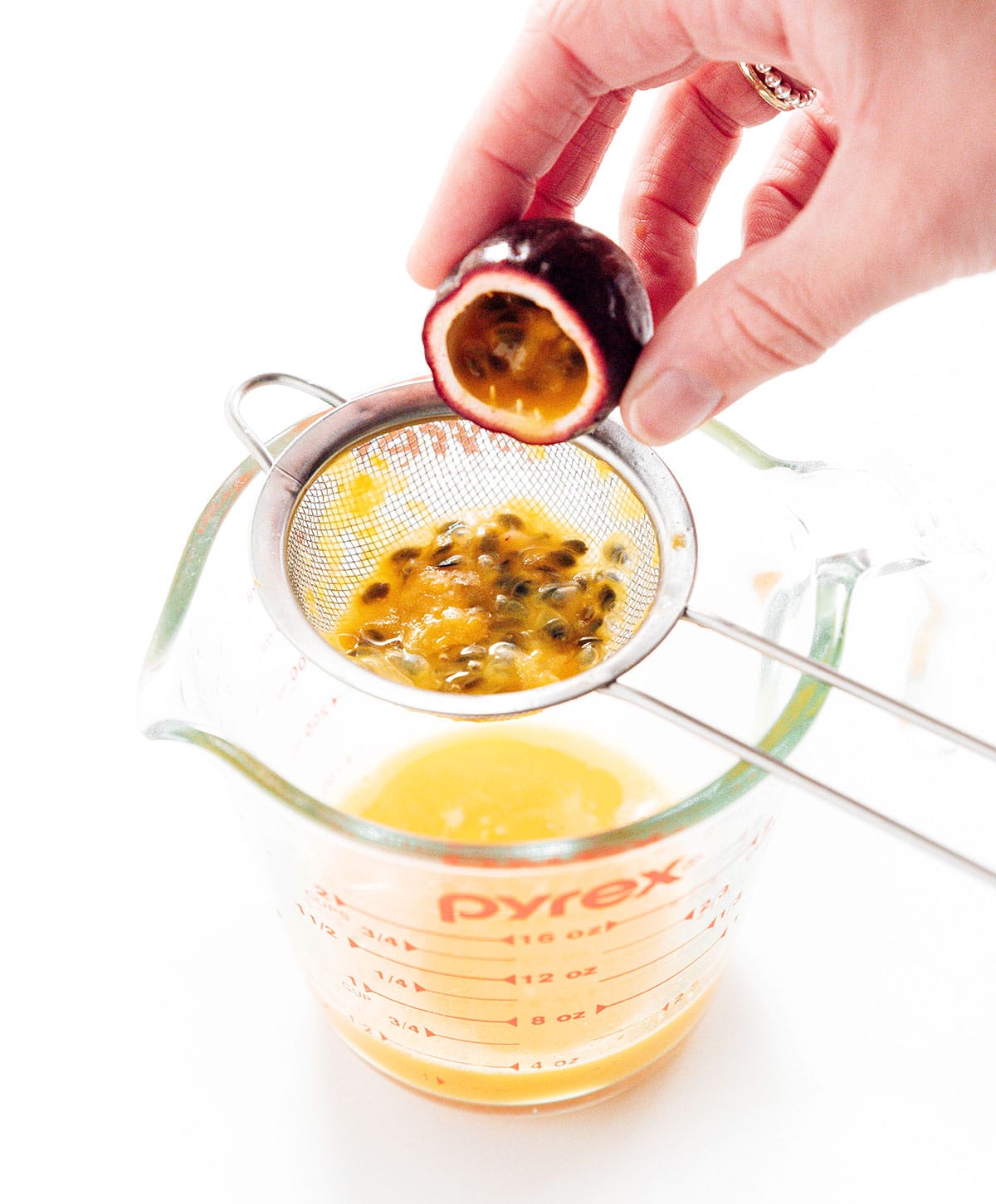 A hand emptying a cut open passion fruit into a small strainer over a measuring cup.