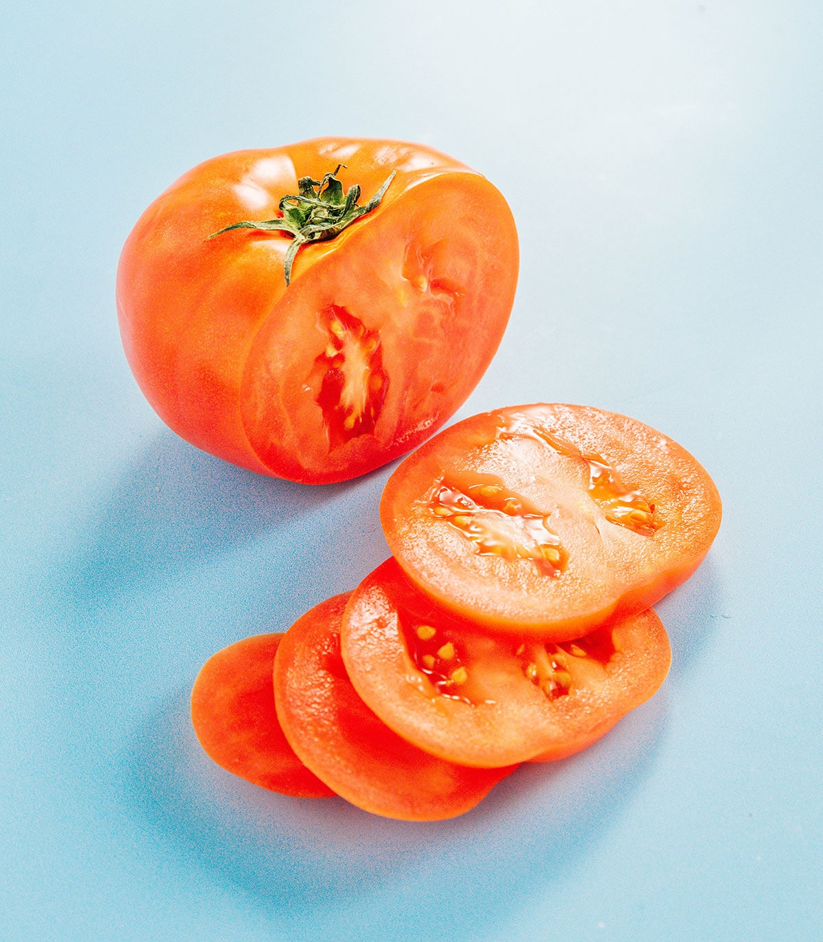 A tomato with half of it cut in slices on a blue surface.
