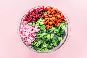 A glass bowl of broccoli salad features chopped red onion, dried cranberries, roasted almonds, diced cucumber, and broccoli florets on a pink background.