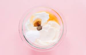 A glass bowl containing ingredients for broccoli salad—sour cream, mayonnaise, sugar, black pepper, and vinegar—viewed from above on a pink background.