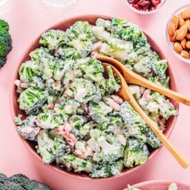 A bowl of broccoli salad with creamy dressing, mixed with chopped nuts and dried cranberries, sits on a pink surface alongside small bowls of almonds and cranberries.