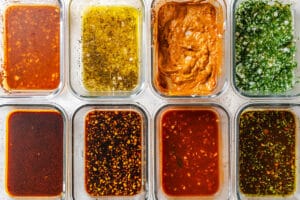 Eight glass containers with different sauces and marinades, perfect for learning how to marinate tempeh, are arranged in two rows of four on a white surface, showcasing a variety of colors and textures.
