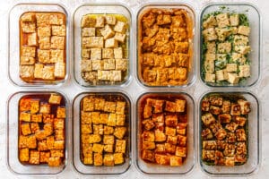 Eight glass containers filled with cubed tempeh, each showcasing how to marinate tempeh in different colorful sauces or seasonings, neatly arranged in two rows on a light surface.