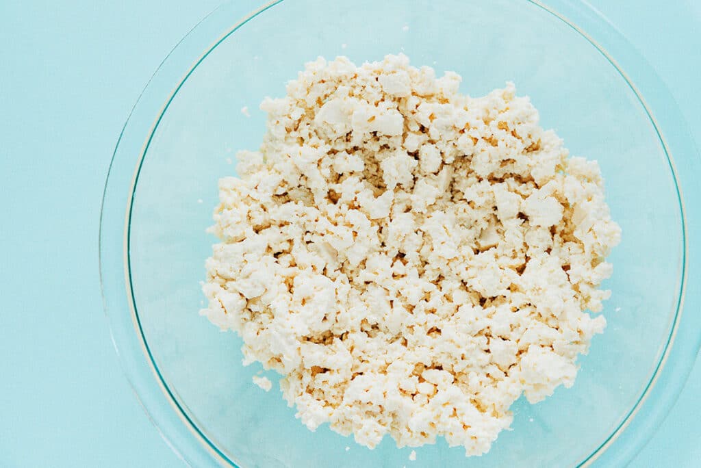 Crumbled tofu in a large glass mixing bowl on a light blue surface.