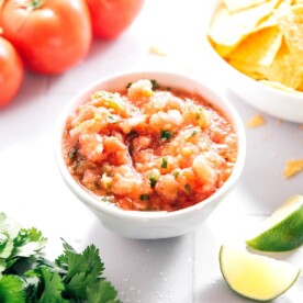 A bowl of vibrant blender salsa sits on a white surface, surrounded by fresh tomatoes, cilantro, a lime wedge, and a bowl of crispy tortilla chips.