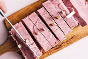 A hand slices pink, frozen loaf pan popsicles with popsicle sticks on a wooden cutting board.