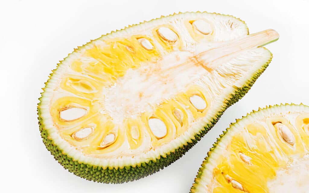 Jackfruit cut in half with the seeds and blubs showing and the bright yellow interior and green bumy exterior.