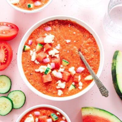 Cold watermelon gazpacho soup in a bowl with a spoon and assorted ingredients around it