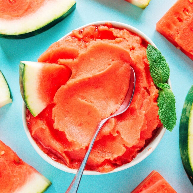 A bowl of watermelon sorbet in a bowl with a spoon surrounded by slices of watermelon on a blue background