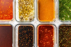 Eight clear containers filled with different sauces, dressings, and tofu marinades—each with varying colors and visible herbs, spices, or oil—are arranged in a neat grid.