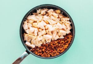 A frying pan containing separated sections of chopped jackfruit and browned crumbled soy chorizo—perfect for creating a jackfruit curry—on a pale green background.