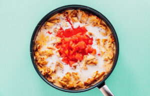 A frying pan containing shredded chicken, coconut milk, and diced tomatoes sits on a light teal background, perfect for a comforting jackfruit curry-inspired dish.