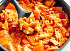A close-up of a pan filled with jackfruit curry, featuring chunks of jackfruit, bell peppers, and tomatoes in a reddish-orange sauce, with a ladle scooping some of the mixture.