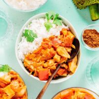 A bowl of white rice with flavorful jackfruit curry, garnished with cilantro, sits on a table alongside extra jackfruit curry bowls, a spice dish, water glasses, and a piece of jackfruit.