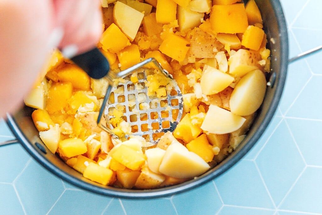 A potato masher being used in a pot of cooked potatoes and rutabaga.