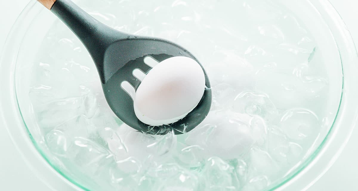 A slotted spoon adding a boiled egg to a bowl of ice water.