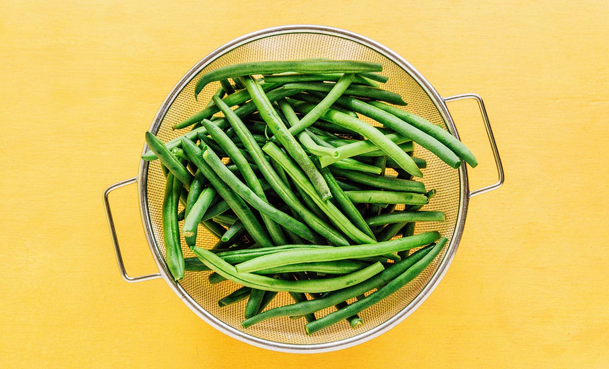 Raw green beans with ends trimmed in a mesh colander.