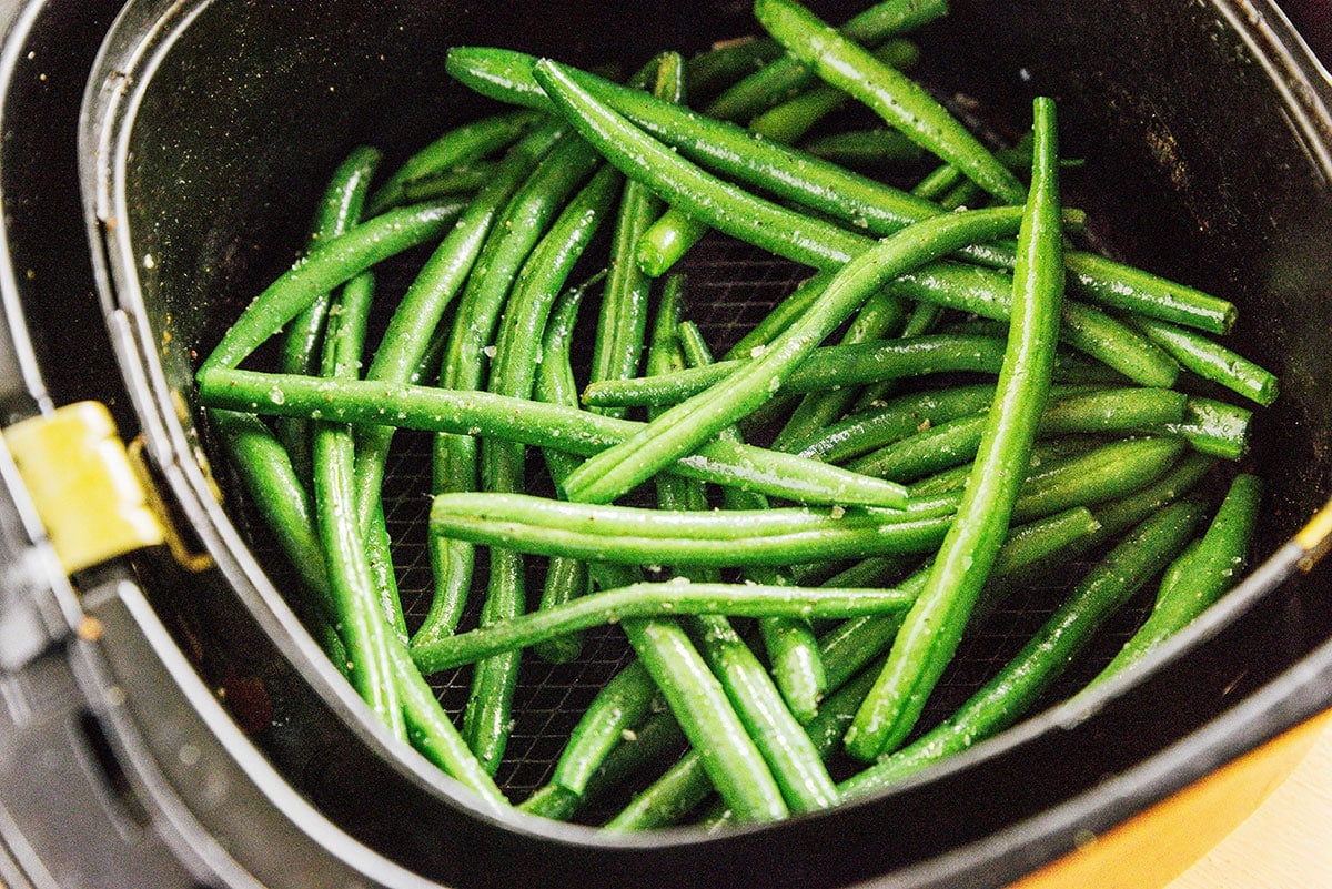 Oiled and seasoned green beans in the basket of an air fryer.