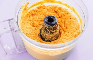 A food processor bowl filled with orange-colored pumpkin hummus, evenly spread around the blade in the center.