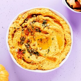A bowl of creamy pumpkin hummus topped with seeds and herbs sits on a light purple surface, surrounded by pita chips and small pumpkins.