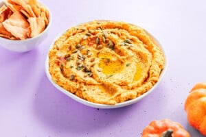 A bowl of pumpkin hummus topped with pumpkin seeds and herbs sits on a light purple surface next to a bowl of pita chips and a small pumpkin.