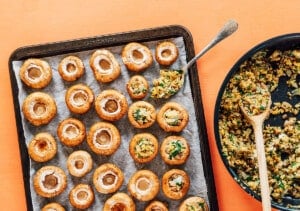 A baking tray with hollowed-out mushrooms on parchment paper, perfect for preparing vegetarian stuffed mushrooms, sits beside a pan of chopped vegetable filling and a spoon.