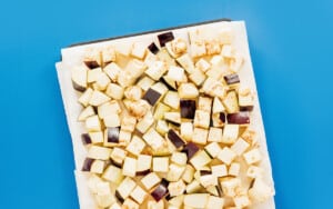 A baking tray lined with parchment paper holds a single layer of cubed raw eggplant, ready to be transformed into delicious air fryer eggplant on a bright blue background.