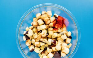 A glass bowl containing chopped air fryer eggplant pieces and assorted spices sits on a blue surface.