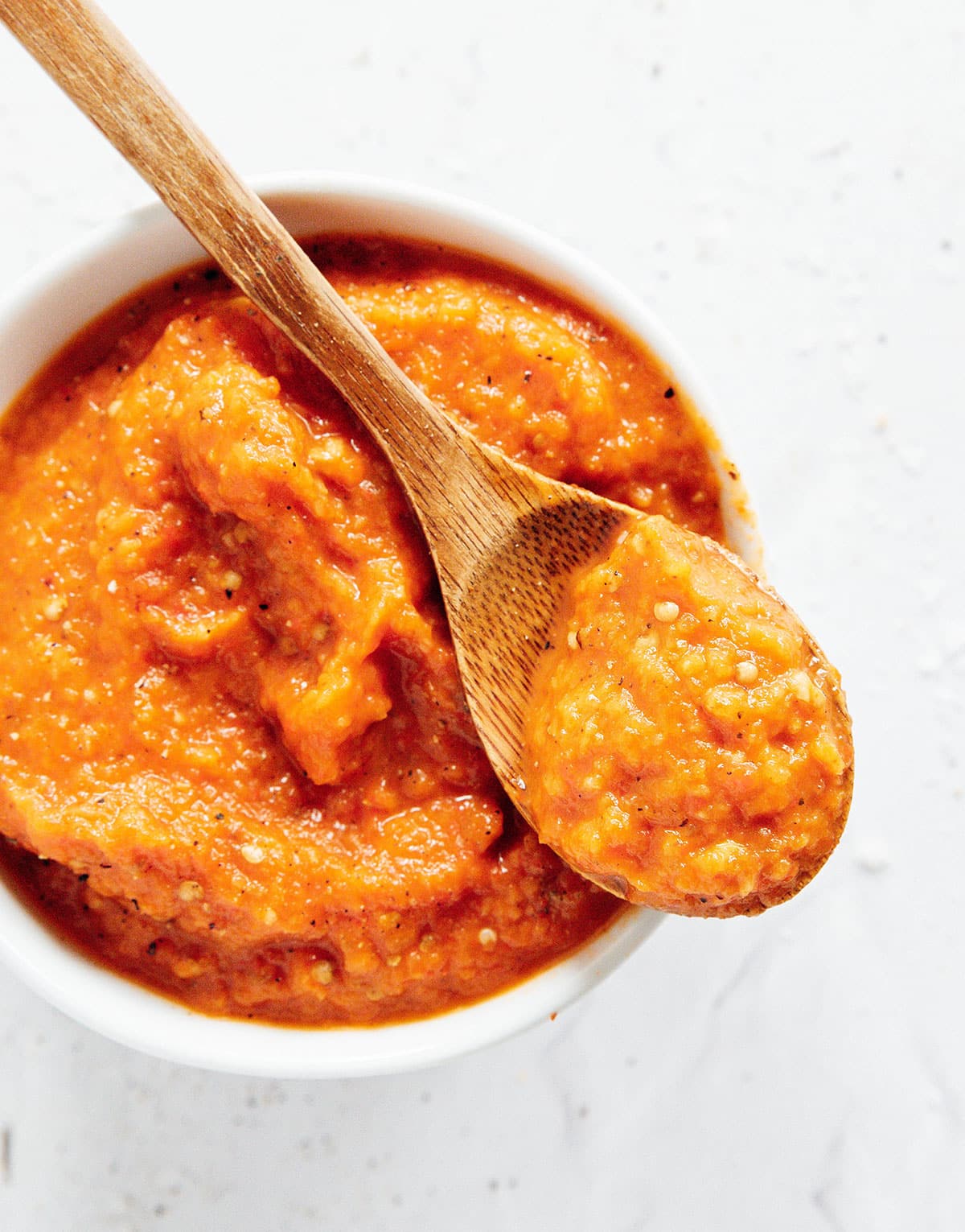 A wooden spoon with a scoop of ajvar sauce from a white bowl.