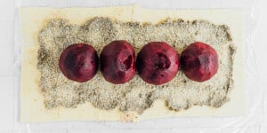 Four peeled beets are placed in a row on a bed of herbs and grains atop a rectangular sheet of pastry dough, ready to be wrapped into a vibrant beet wellington.