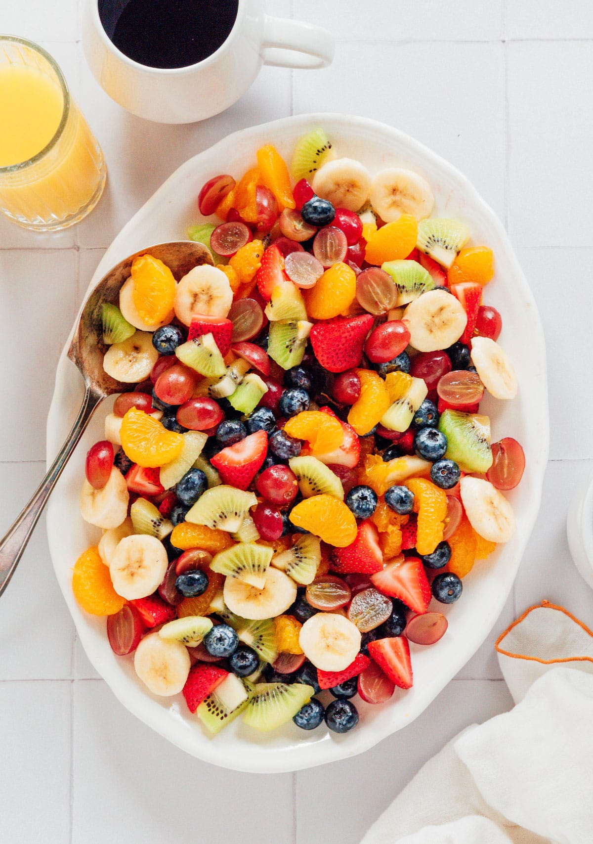 Breakfast fruit salad in a large white bowl with a glass of orange juice and coffee mug nearby.