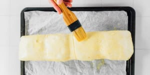 A hand uses a pastry brush to apply egg wash to a large, rectangular beet wellington dough on a parchment-lined baking tray.