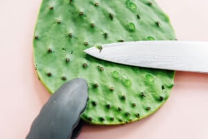 A knife is being used to remove spines from a green cactus pad on a light surface, an essential first step in learning how to cook nopales.