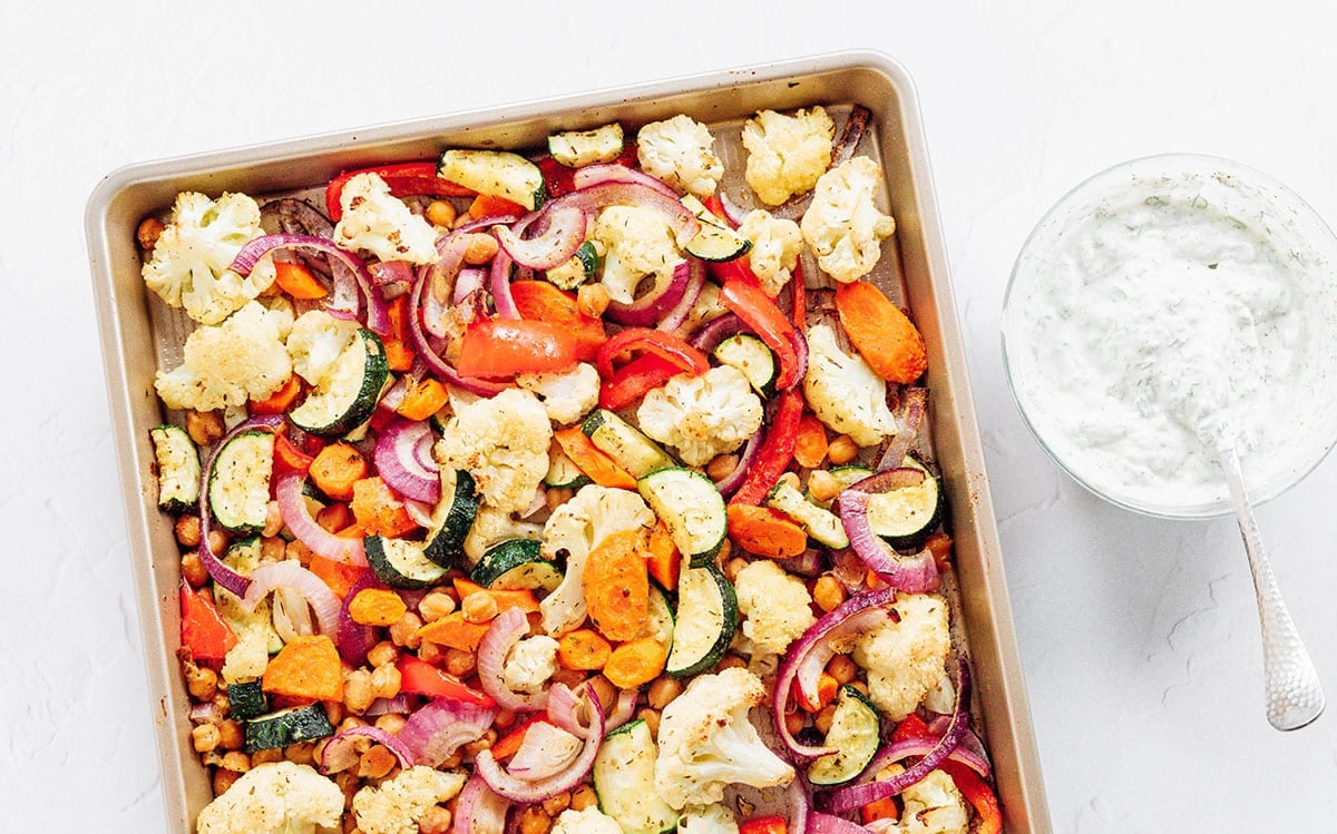 A sheet pan of roasted Mediterranean flavored veggies with a bowl of dill yogurt sauce.