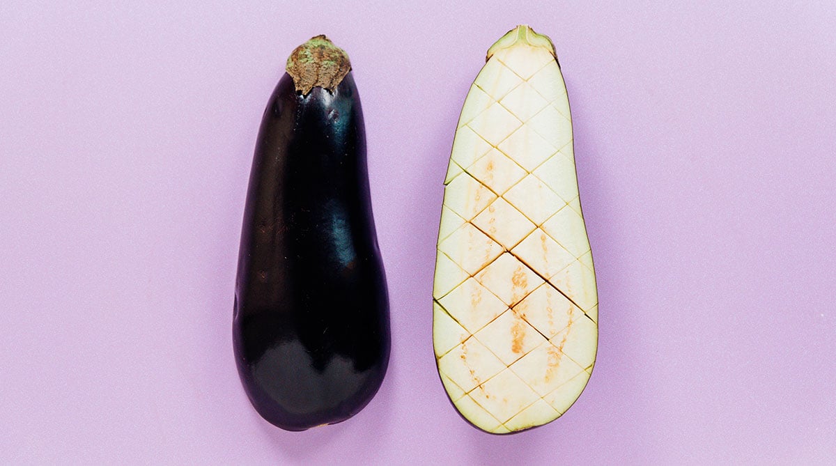 A whole eggplant next to a halved one that has crosshatches in the flesh.