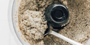 A close-up of a food processor containing a coarse, blended beet wellington mixture being scooped out with a metal spoon.