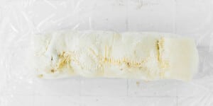 A rolled and sealed log of raw pastry dough, prepped for beet wellington, rests on clear plastic wrap, with visible fork marks along the seam.