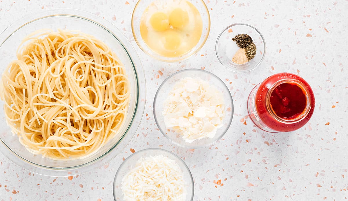 Ingredients for spaghetti pizza in bowls on a white surface.