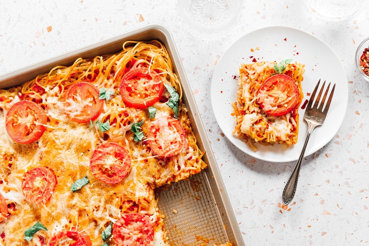 A fork and serving of spaghetti pizza on a small white plate next to a baking dish.