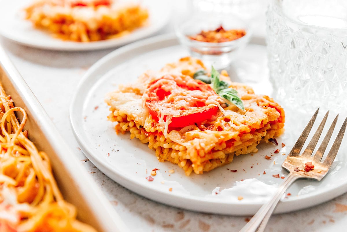 Side view of a slice of spaghetti pizza with sliced tomato on a plate.
