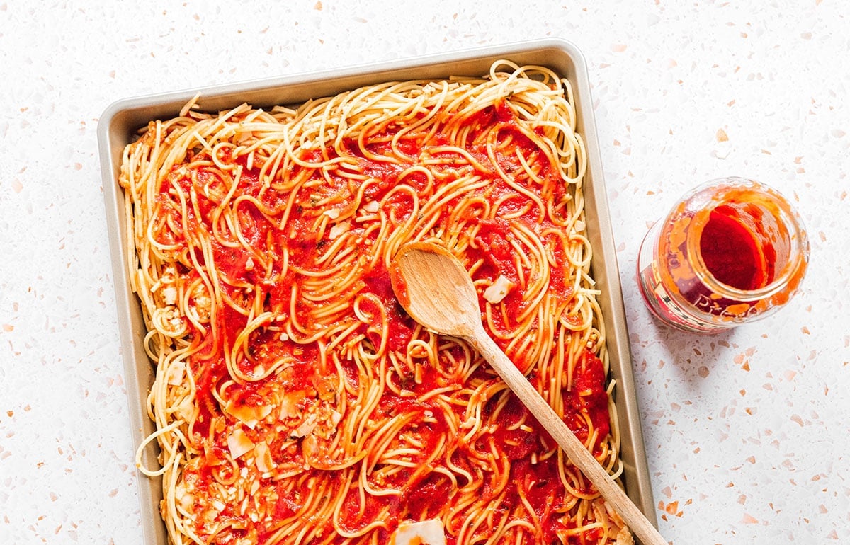 A wooden spoon spreading pasta sauce over a tray of spaghetti.