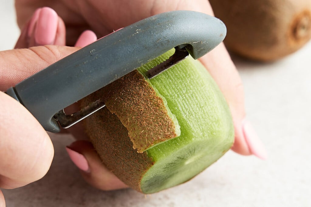 Peeling Option 2: You can also use a vegetable peeler to remove the skin. Hold the kiwi in the palm of your hand or against the counter. Using vertical motions, run the peeler all around the kiwi until the skin is gone.