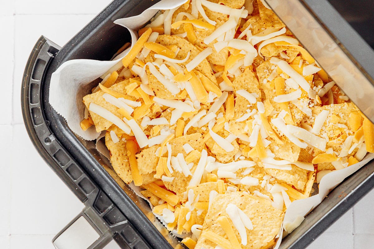 Tortilla chips and shredded cheese layered in the basket of an air fryer.