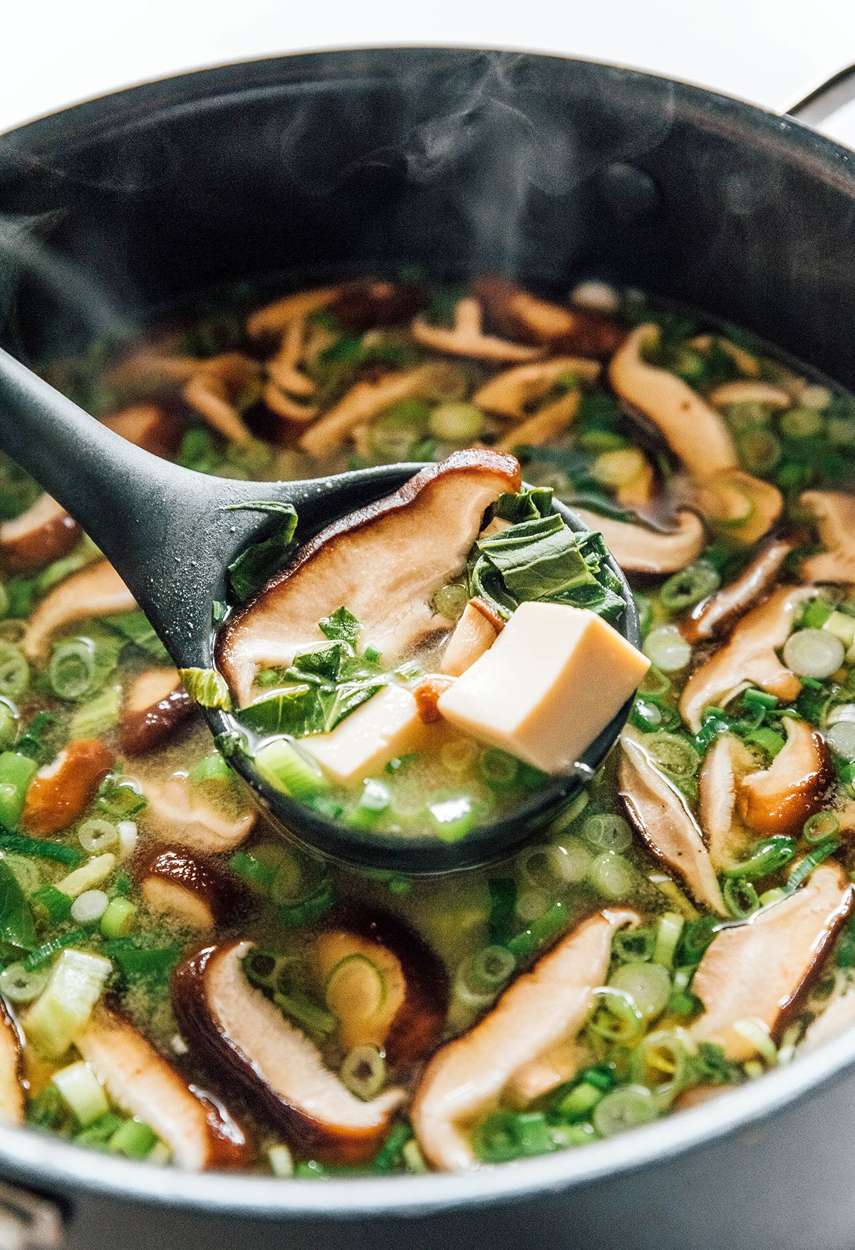 A ladle scooping out vegan miso soup with bits of bok choy, mushrooms, tofu, and green onions.