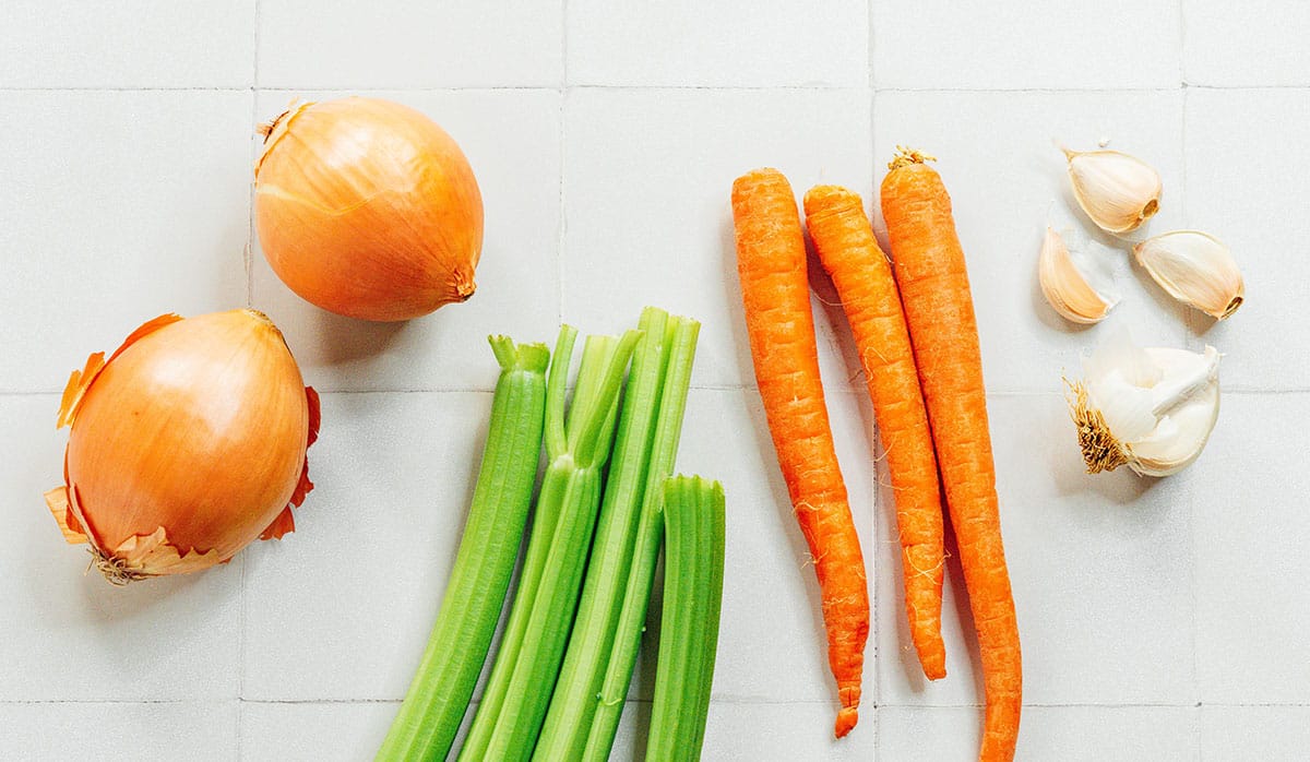 Carrots, celery, carrots, and garlic set on a white counter before chopping. 