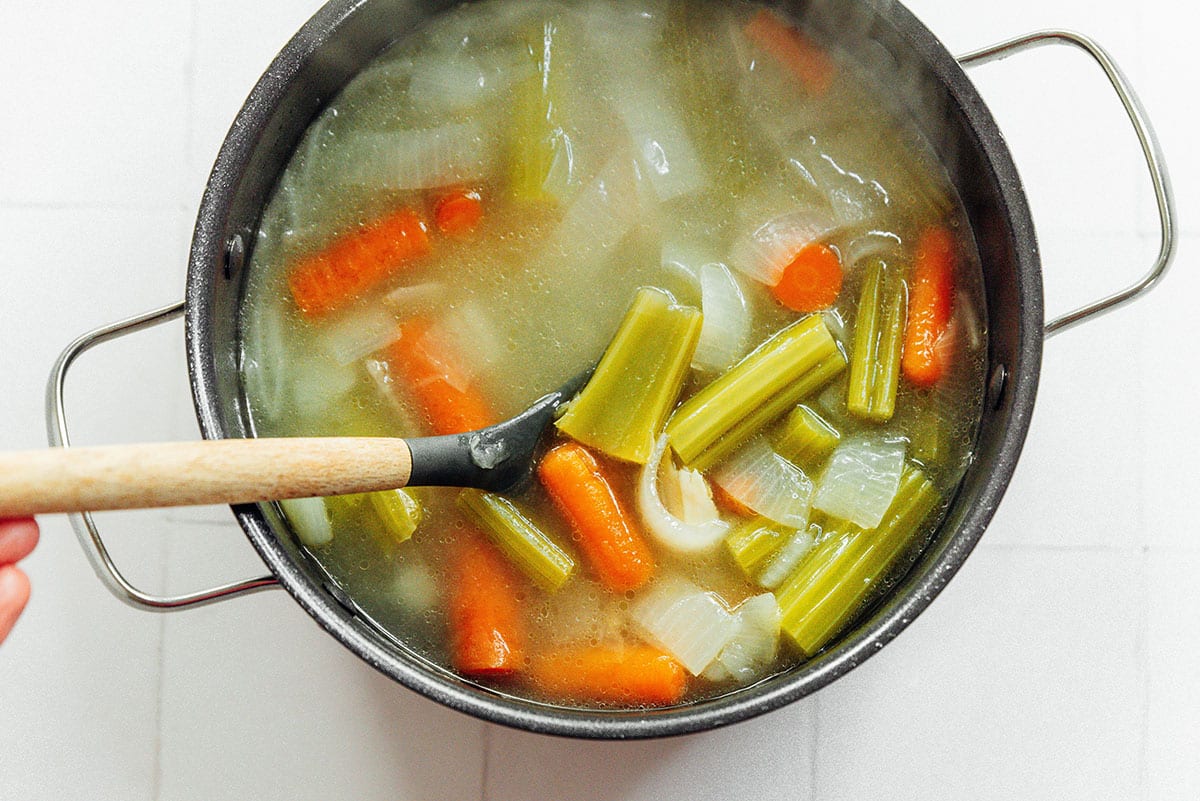 Steaming broth filled with fresh vegetables in a large black pot on a white background. A wooden spoon rests in the pot.