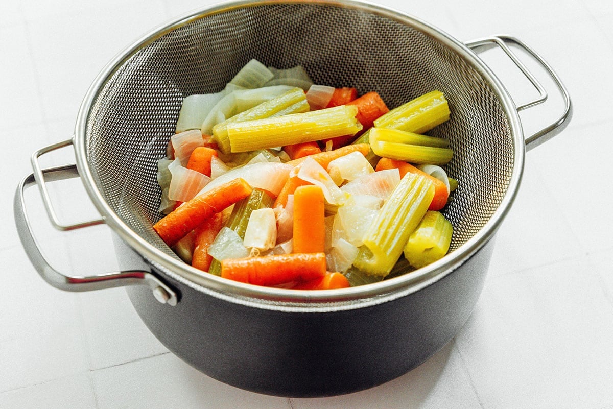 Cooked fresh vegetables in a large silver strainer, set on top of a large black pot. White background.